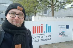 Mgr Wojciech Wydmański, doktorant UKSW, podczas wizyty badawczej na Uniwersytecie w Luxemburgu, ubrany w czapkę i szalik, pozuje do zdjęcia przed budynkiem z logo Uniwersytetu.