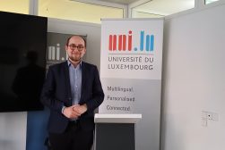 Mgr Wojciech Wydmański, doktorant UKSW, podczas wizyty badawczej na Uniwersytecie w Luxemburgu, ubrany w garnitur, pozuje do zdjęcia przed logo Uniwersytetu.