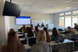 Dr Magdalena Maksymiuk, w sali wykładowej, podczas prowadzenia zajęć dla studentów, w ramach stażu na Uniwersytecie Wileńskim. Na pierwszym planie słuchacze, w tle Pani dr i badaczka z Uniwersytetu Wileńskiego prowadzą wykład. Na ekranie wyświetlona prezentacja.