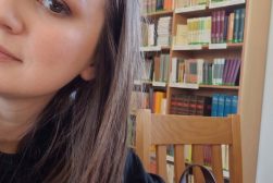 Dr Magdalena Maksymiuk, w bibliotece w trakcie stażu na Uniwersytecie Wileńskim. Na pierwszym planie Pani dr., w tle regał z książkami.