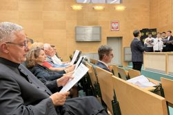 Publiczność śpiewa pieśni patriotyczne z rozdanymi tekstami, podczas gdy dyrygent prowadzi występ chóru w sali Wydziału Prawa i Administracji UKSW.