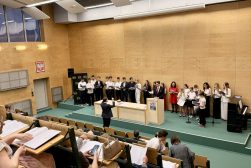 Dyrygent prowadzi chór podczas koncertu pieśni patriotycznych w sali Wydziału Prawa i Administracji UKSW. Na pierwszym planie widoczna widownia spiewająca wraz z churem