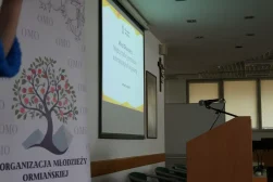 Na pierwszym planie pusta mównica i logo Organizacji młodzieży ormiańskiej w Polsce. W tle ekran, na którym wyświetlona jest prezentacja. Na dalszym planie portret Jana Pawła Drugiego i Krzyż.