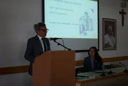Uczestnik Konferencji wygłaszajacy wykład podczas konferencji naukowej „Dziedzictwo Alfreda Ohanowicza”. Prof. Stoi za mównicą. Przy stole prezydialnym siedzi kobieta - organizatorka konferencji. W tle ekran, na którym wyświetlona jest prezentacja. Na dalszym planie portret Jana Pawła Drugiego i Krzyż.
