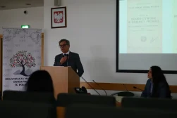 Uczestnik Konferencji wygłaszajacy wykład podczas konferencji naukowej „Dziedzictwo Alfreda Ohanowicza”. Prof. Stoi za mównicą. Przy stole prezydialnym siedzi kobieta - organizatorka konferencji. W tle ekran, na którym wyświetlona jest prezentacja.