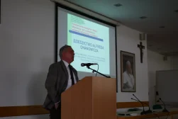 Prof. Dariusz Fuchs wygłaszajacy wykład podczas konferencji naukowej „Dziedzictwo Alfreda Ohanowicza”. Prof. Stoi za mównicą. W tle ekran, na którym wyświetlona jest prezentacja. Na dalszym planie portret Jana Pawła Drugiego i Krzyż.
