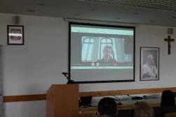 Uczestniczka Konferencji wygłaszajacy wykład za pośrednictwem wideo rozmowy podczas konferencji naukowej „Dziedzictwo Alfreda Ohanowicza”. Wideorozmowa wyświetlana na ekranie. W tle Godło Polski oraz portret Jana Pawła drugiego i Krzyż.