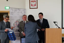 Prof. Dariusz Fuchs wraz z dwoma mężczyznami, uczestnikami konferencji naukowej „Dziedzictwo Alfreda Ohanowicza”, odbierający od organizatorki nagrody. Wszyscy są elegancko ubrani. Pan Prof. i pozostali mężczyźni trzymają niebiesko-granatowe torby prezentowe.