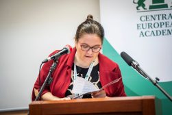 Prof. Aleksandra Syryt w czerwonej marynarce odczytuje wystąpienie przy mównicy, między dwoma mikrofonami, na tle baneru Central European Academy