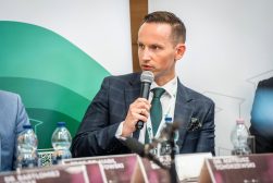 Mateusz Kaczorowski zabiera głos podczas panelu, siedząc przy stole z mikrofonem, wizytówkami i butelkami wody