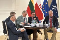 Uczestnicy panelu dyskusyjnego siedzą przy stole; jeden z mężczyzn mówi do mikrofonu, a w tle widoczne są flagi Litwy, Polski, Unii Europejskiej i NATO
